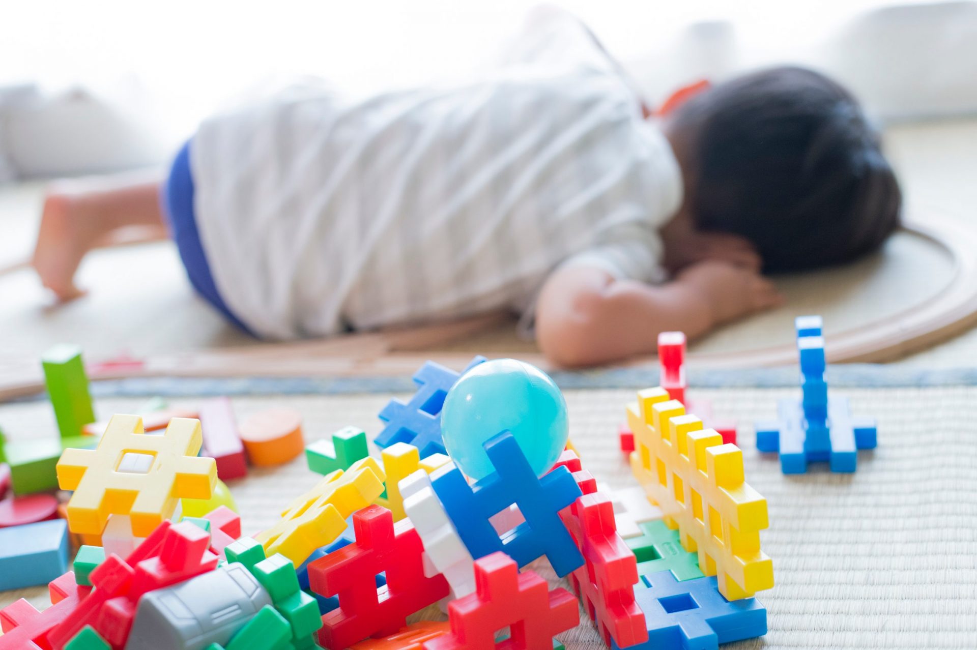 Das Bild zeigt ein kleines Kind, wahrscheinlich ein Kleinkind, das mit dem Gesicht nach unten auf einer hellen Matte liegt. Der Körper des Kindes ist unscharf, was darauf hindeutet, dass der Fokus auf den Spielzeugen liegt. Das Kind trägt ein helles, kurzärmeliges Hemd und scheinbar blaue Shorts. Sein dunkles Haar ist sichtbar. Die Körperhaltung des Kindes lässt vermuten, dass es schläft oder sich ausruht. Im Vordergrund ist ein Stapel bunter Plastikbausteine zu sehen, die scharf abgegrenzt sind. Diese Blöcke haben verschiedene Formen und Größen, wobei die meisten aus ineinandergreifenden „+“-förmigen Teilen in den Farben Rot, Gelb, Blau und Grün bestehen. Es gibt auch einige andere Formen und Farben sowie eine hellblaue Plastikkugel, die sich zwischen den Blöcken befindet. Die Blöcke scheinen eine Art Konstruktionsspielzeug zu sein. Die Matte unter dem Kind und den Spielzeugen scheint ein helles, gewebtes Material mit einer feinen Struktur zu sein. Der Hintergrund ist unscharf, deutet aber auf einen hell erleuchteten Raum hin, möglicherweise ein Spiel- oder Schlafzimmer. Die Gesamtatmosphäre ist ruhig und friedlich, was auf einen Moment der Ruhe nach dem Spielen hindeutet.